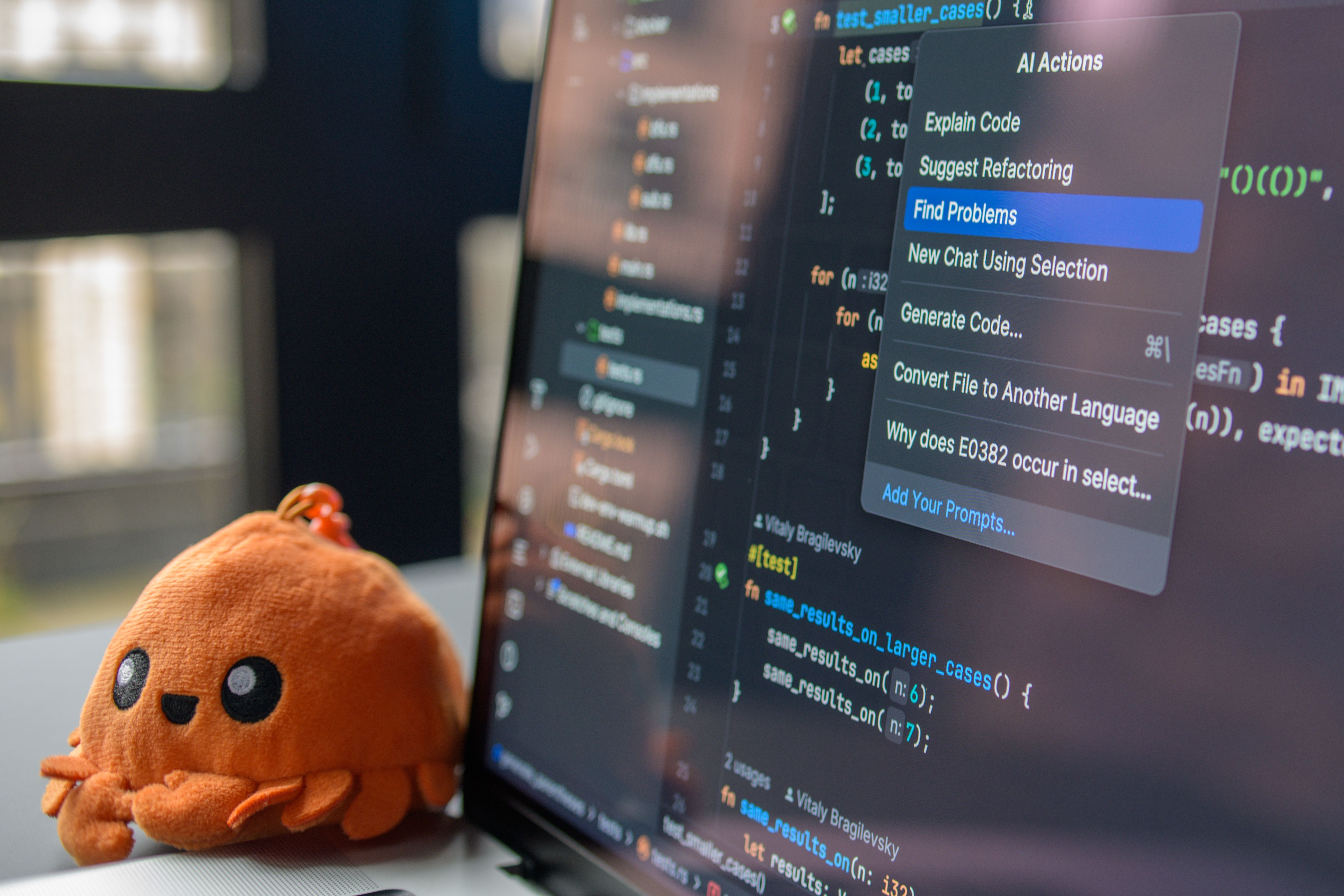 A plush crab toy beside a monitor showing a code editor, an assistant menu open over the file listing actions like Explain Code and Find Problems.